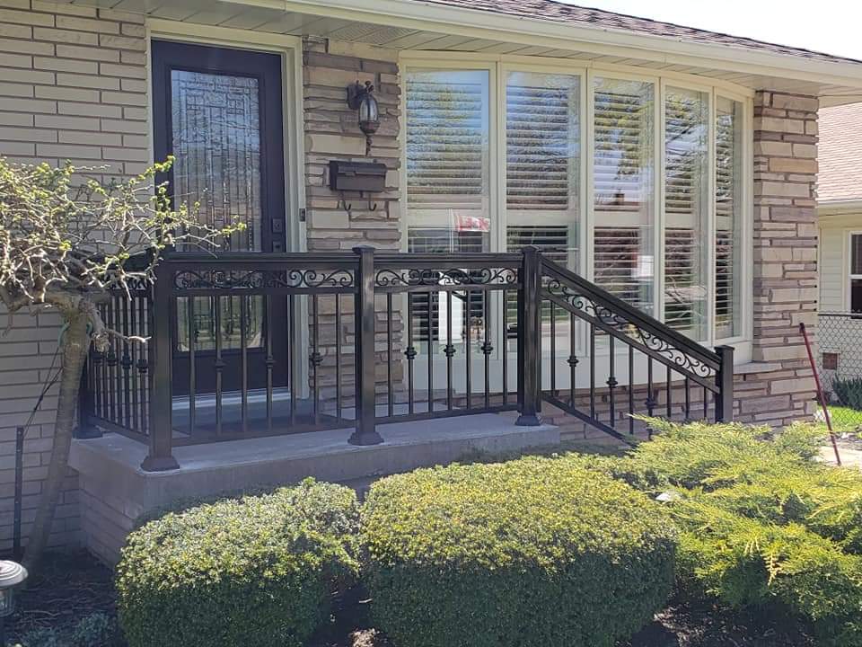 Black ornamental metal railing on a front porch with stone siding and green bushes.