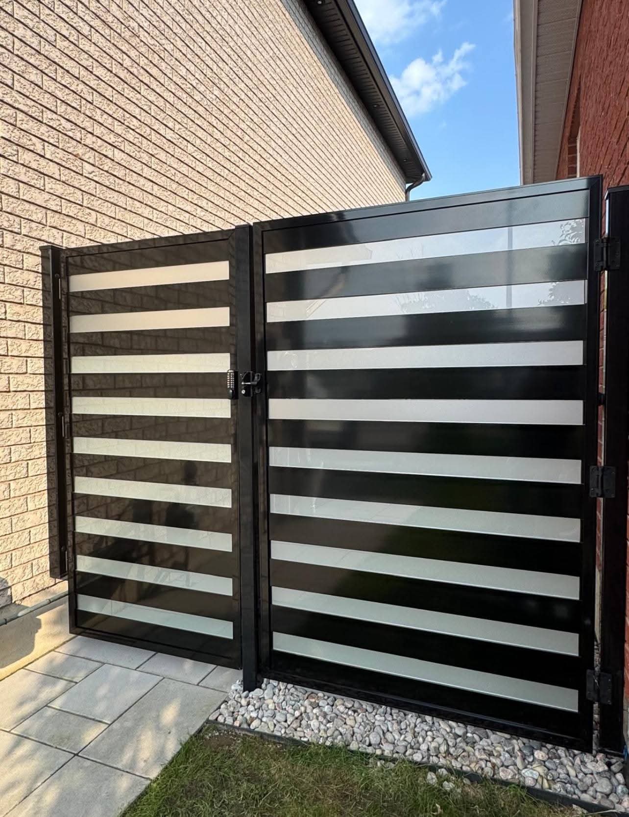 Modern black metal gate with horizontal frosted glass panels between two residential brick houses.