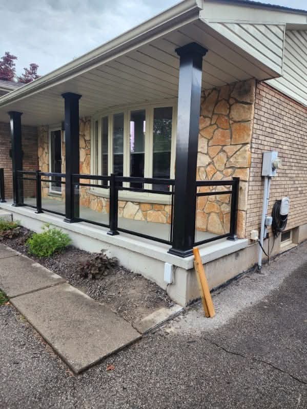 Modern front porch featuring black support columns, glass panel railings, and a tan stone facade.
