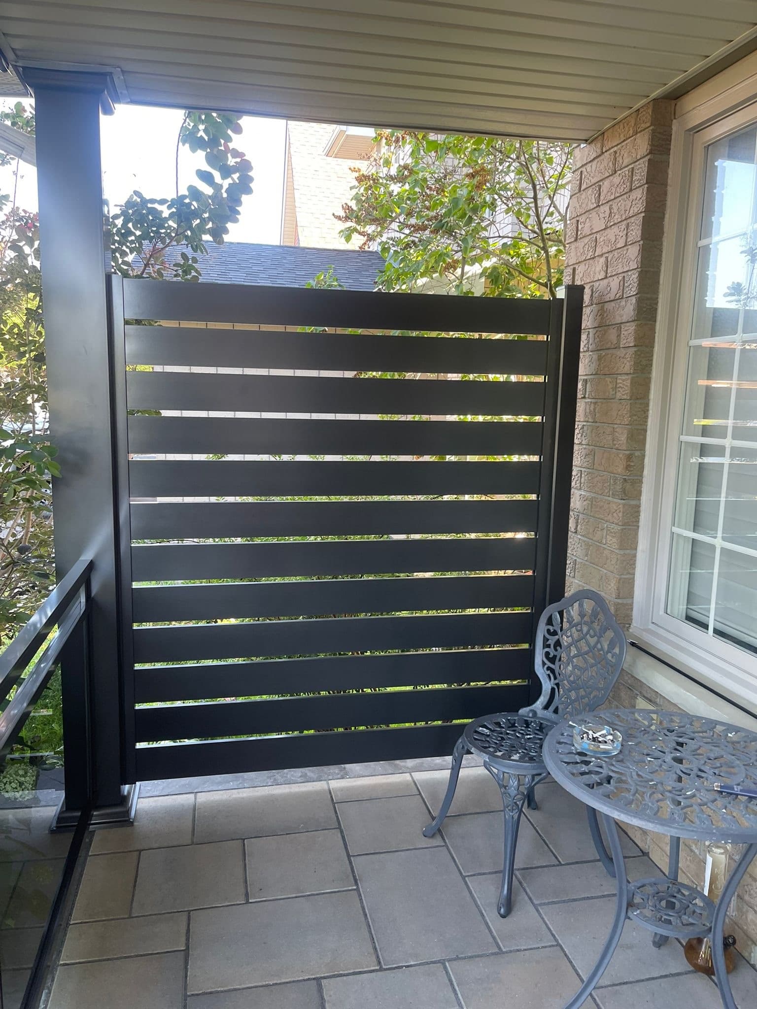 Black horizontal slat privacy screen on a stone patio next to grey bistro furniture.