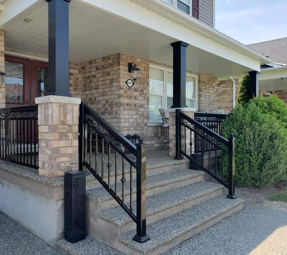 Aggregate steps with black decorative metal railings leading to a brick home's covered front porch.